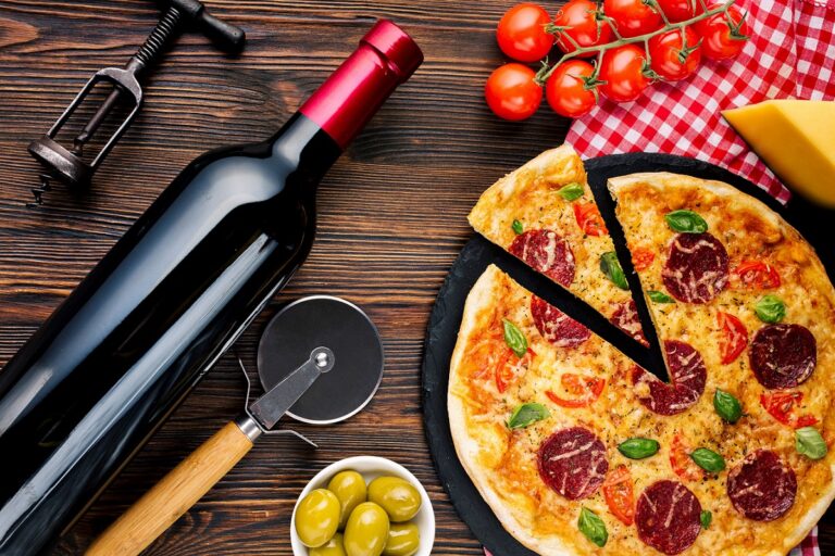Overhead view of bottle of wine and pizza with pizza cutter, olives, and tomatoes.