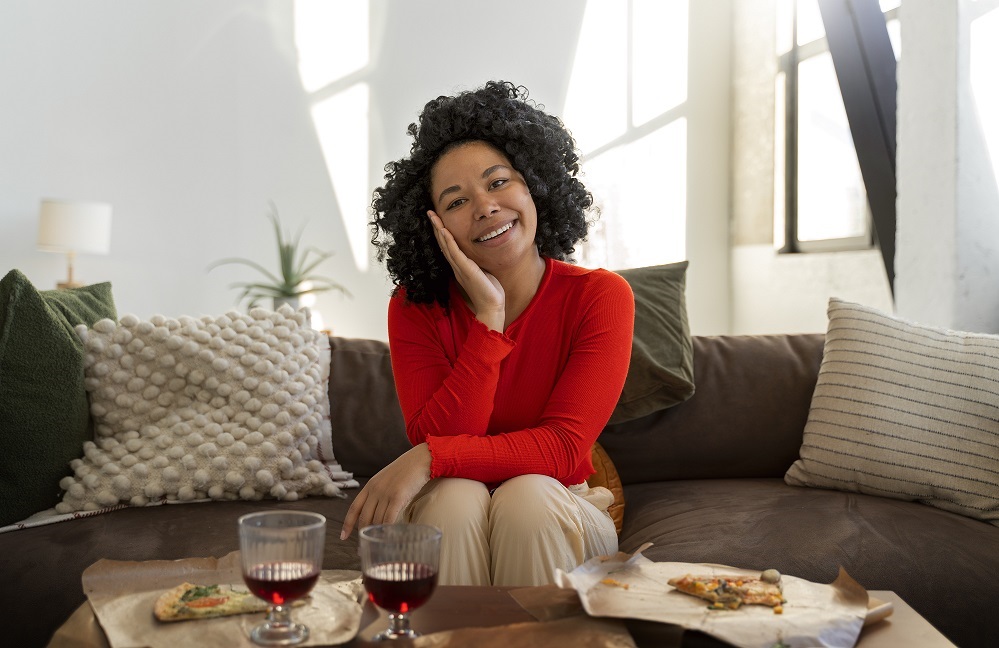 Photo of Black woman sitting on a sofa with glasses of wine and pizza on a table in front of her illustrates blog "What Are Diverse Wine Brands?"
