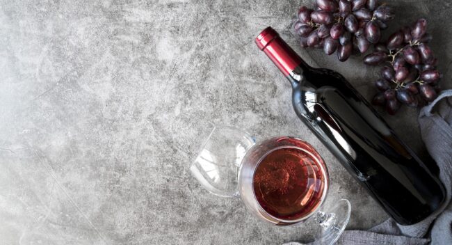 red-table-wine-fp Grapes, bottle of red wine, and glass of wine set against gray background.