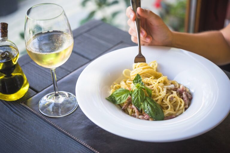 Person holding fork with plate of pasta and glass of wine on the table.